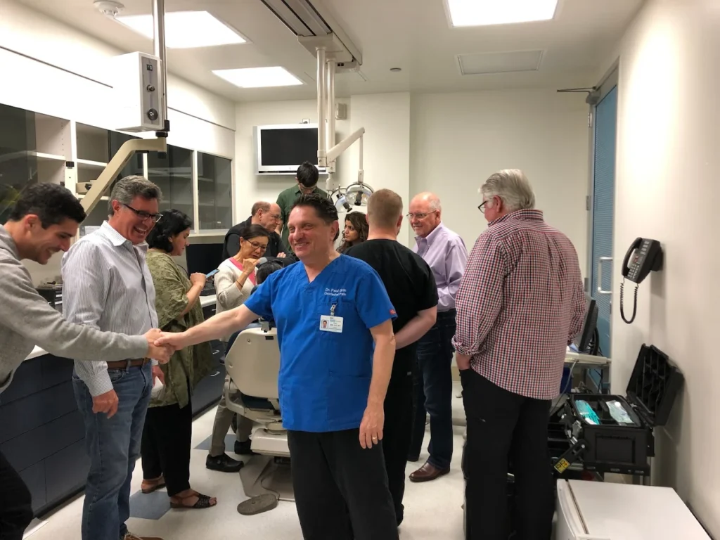 A group of people stand and interact in a medical examination room; one person in scrubs shakes hands with another individual.