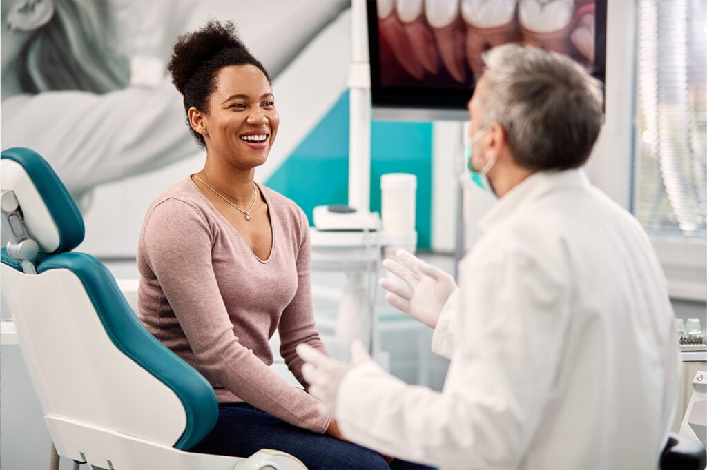 A woman sits in a dental chair, smiling and talking with a dentist who is speaking to her in a brightly lit dental office.