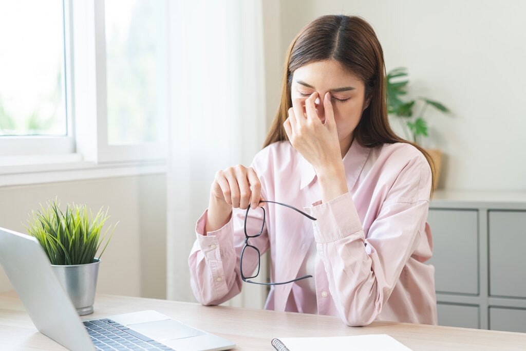 Woman sitting at a desk with a laptop, holding her glasses and pinching the bridge of her nose, appearing tired or stressed.