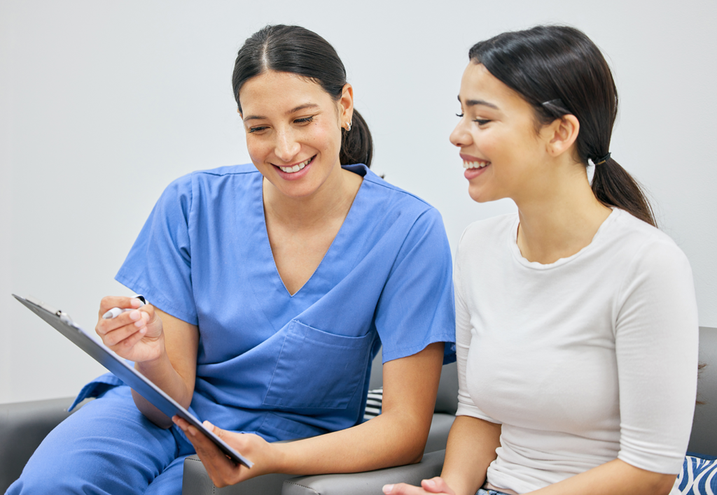 A healthcare professional in blue scrubs discusses notes on a clipboard with a smiling woman in a white top, both seated indoors.