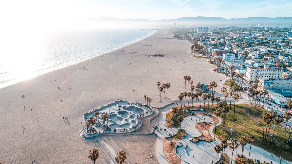Aerial view of a sandy beach with a skatepark, palm trees, and nearby buildings along the coastline under a clear sky.