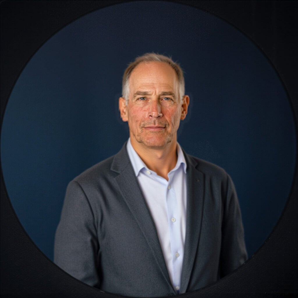 A middle-aged man in a gray suit and white shirt poses against a dark blue background, looking directly at the camera.