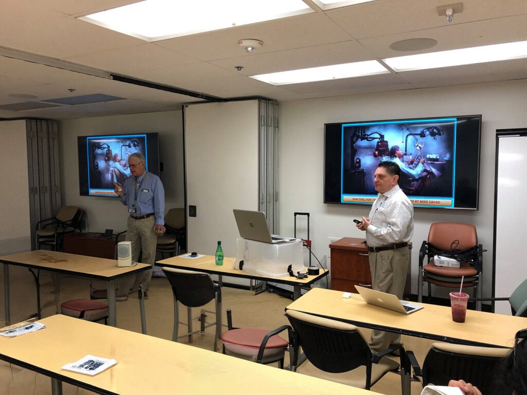 Two men stand at the front of a classroom giving a presentation with slides showing a hospital scene; tables, chairs, and laptops are visible in the room.