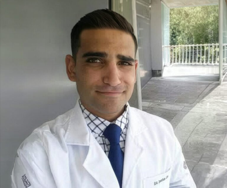 A man wearing a white lab coat, checked shirt, and blue tie stands in front of a modern building with large windows and greenery visible outside.