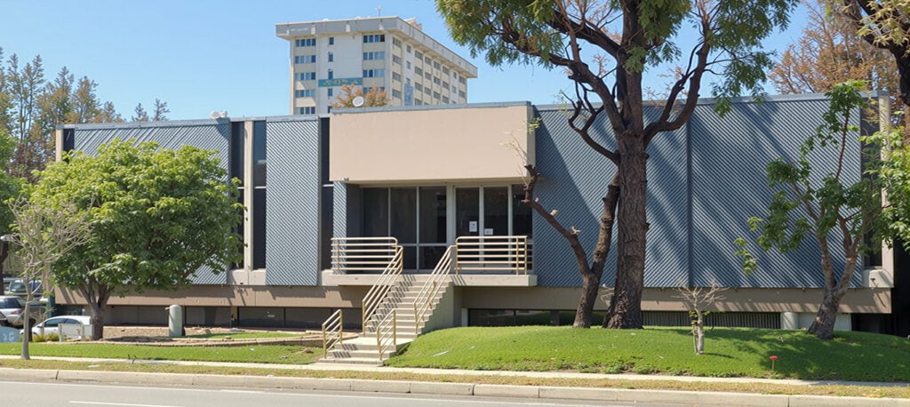 A two-story office building with a flat facade, front stairs, metal railings, and surrounding trees, with a taller building visible in the background.