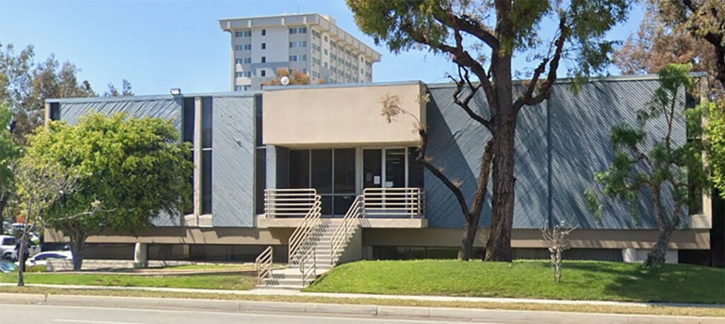 A two-story office building with a flat roof, gray exterior, and front steps leading to the main entrance, situated on a grassy lot with trees; a tall apartment building is visible behind it.