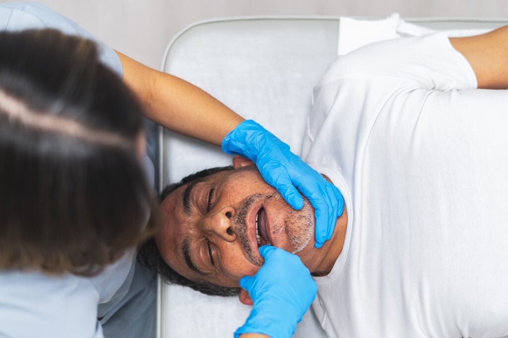 A person lying down receives a mouth examination from a healthcare worker wearing blue gloves.