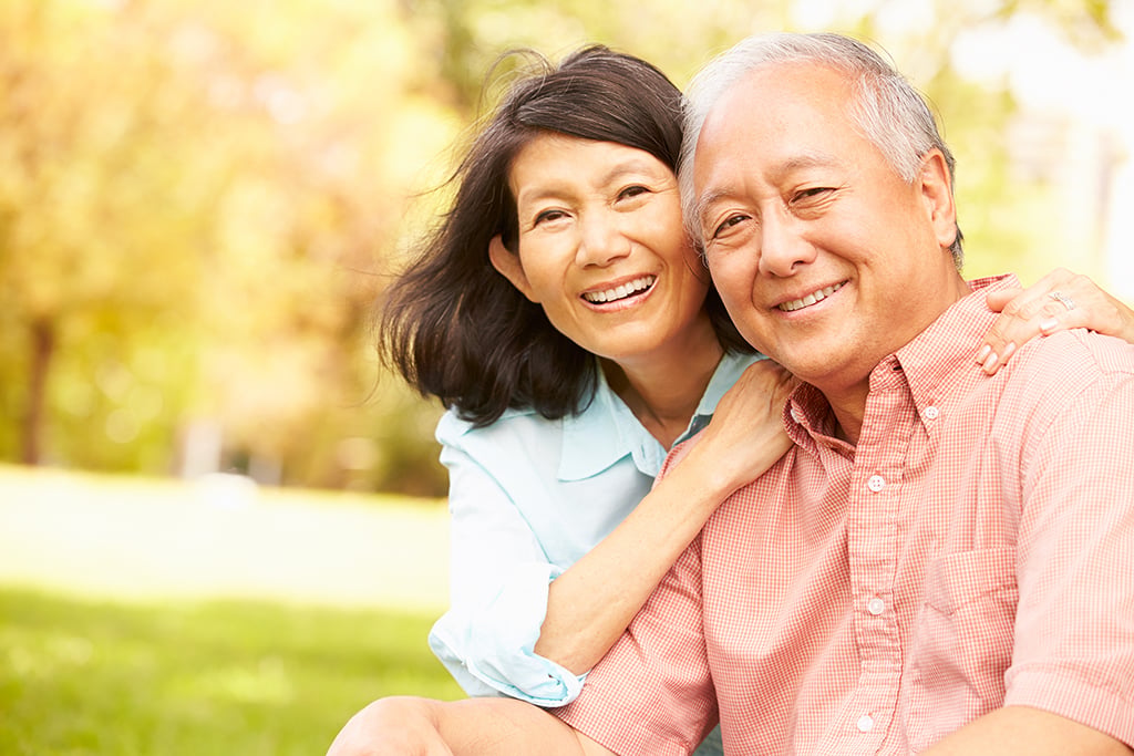 Smiling older couple sitting outdoors in a park, with the woman embracing the man from behind on a sunny day.