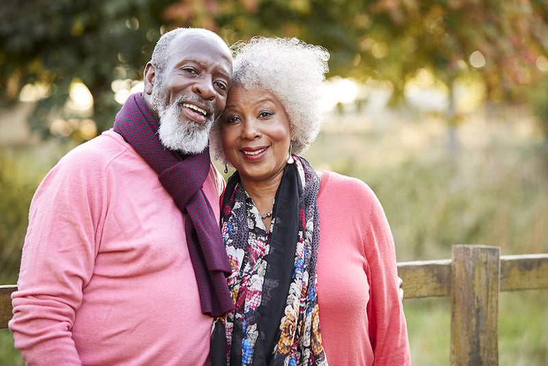 An older couple stands close together outdoors, both smiling. They wear pink sweaters and scarves, with trees and a wooden fence in the background.