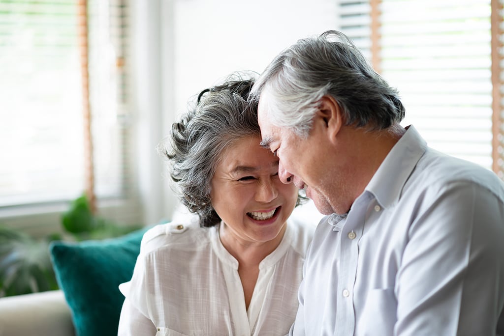 An older couple sits closely together on a sofa, smiling and touching foreheads in a bright room with window blinds in the background.