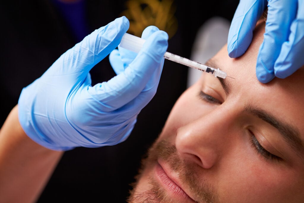 A person receives a cosmetic injection in the forehead while lying down, administered by someone wearing blue medical gloves.