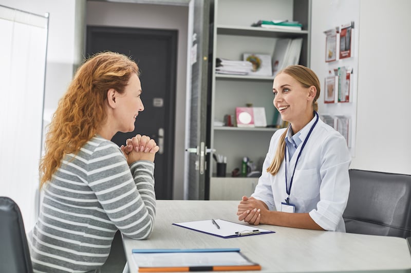 A woman sits across from a healthcare professional in an office, engaged in conversation with a clipboard and pen on the desk between them.