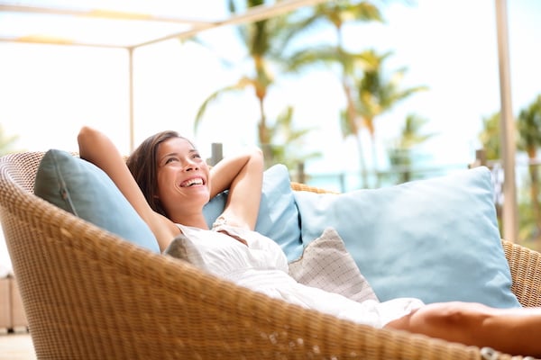 Woman in a white dress relaxing and smiling on a wicker outdoor sofa with blue cushions, palm trees and a bright sky in the background.