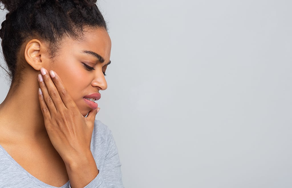 A woman with a pained expression touches her cheek, appearing to experience discomfort or pain in her jaw or mouth.