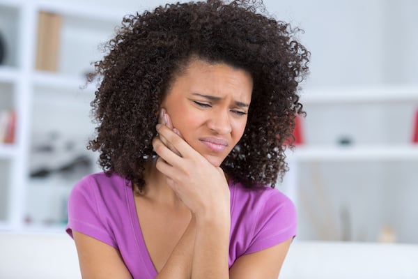 A woman in a purple shirt holds her cheek and appears to be in pain, possibly experiencing a toothache or jaw discomfort.