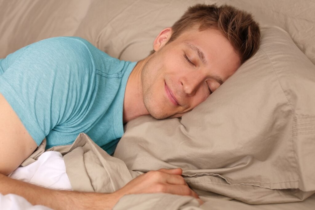 A man with short hair is lying in bed, sleeping on his side with a slight smile, resting his head on a beige pillow.
