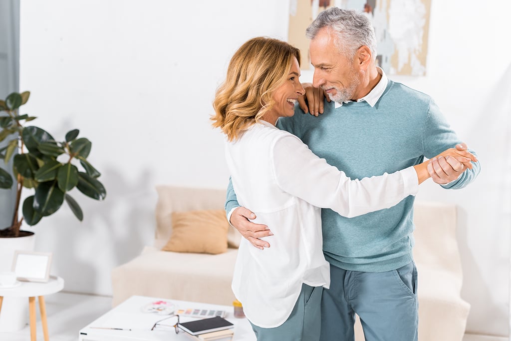 A middle-aged couple dances together in a bright living room, smiling and making eye contact.