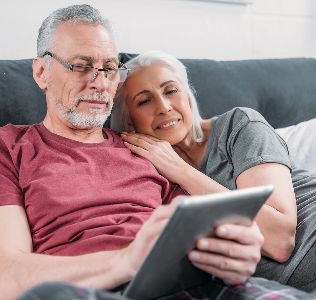 An older man and woman sit on a couch together; the man holds a tablet while the woman leans on his shoulder and smiles.