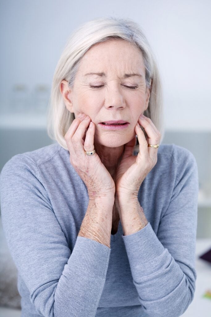 Older woman with closed eyes holding her jaw with both hands, appearing to be in pain or discomfort.