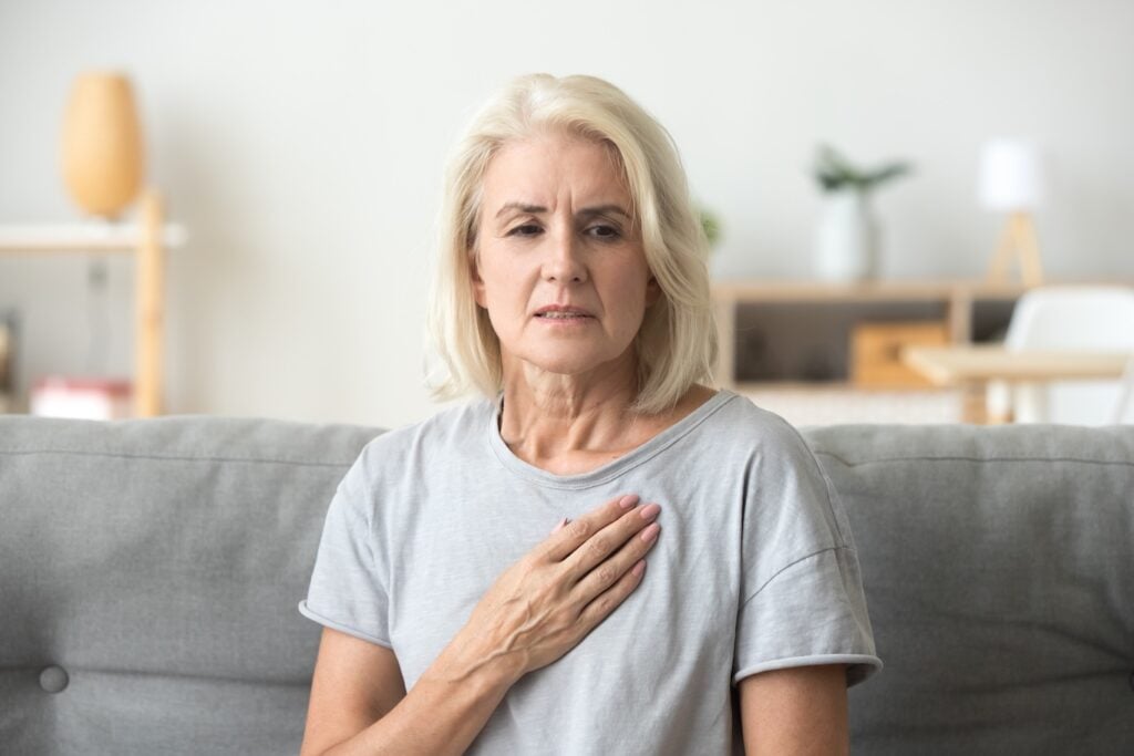 An older woman sits on a couch with her hand on her chest, looking concerned.