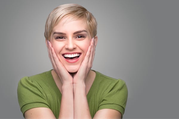 A person with short blond hair, wearing a green shirt, smiles widely with both hands on their cheeks against a plain gray background.