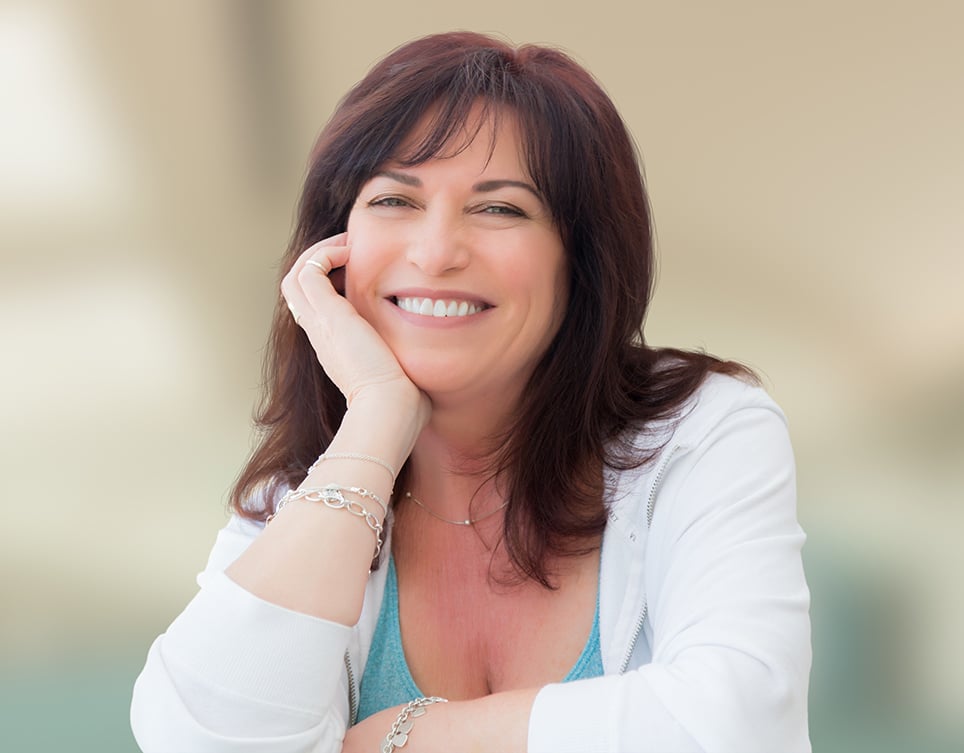A woman with shoulder-length brown hair smiles at the camera, resting her head on her hand. She is wearing a white top and silver bracelets. The background is softly blurred.
