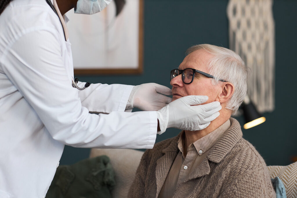 A healthcare professional wearing gloves examines the neck of an older man who is sitting and wearing glasses and a sweater.