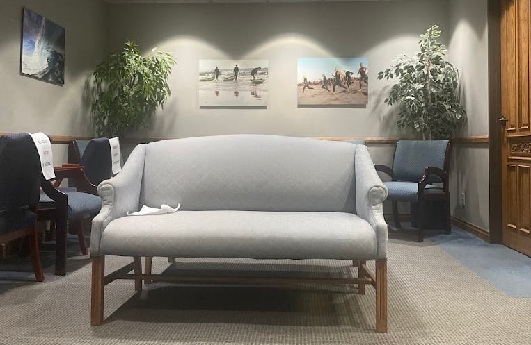 A gray upholstered sofa sits in the center of a waiting room with blue chairs, two potted plants, and three beach-themed photos on the wall behind it.
