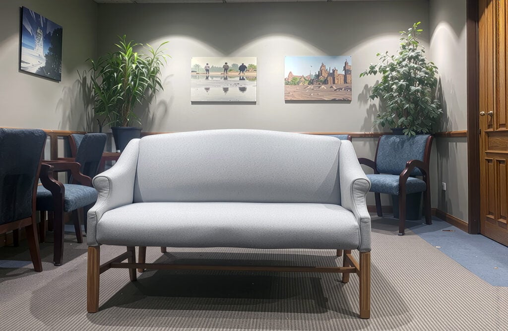 A light gray upholstered sofa is positioned in a waiting area with blue chairs, green plants, and framed artwork on the walls.