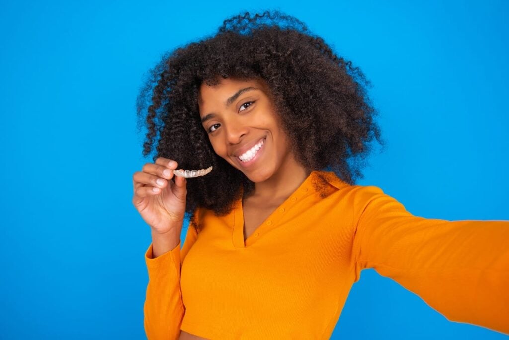 Woman with curly hair smiling and holding a clear dental aligner, wearing an orange top, standing against a solid blue background.